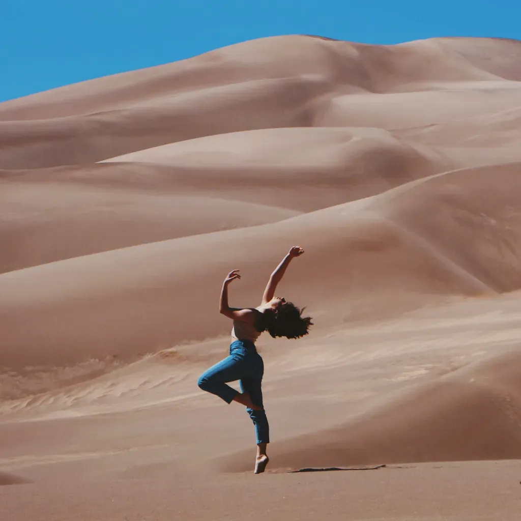 woman standing on right foot and raising hands at the bottom of sand dunes during daytime